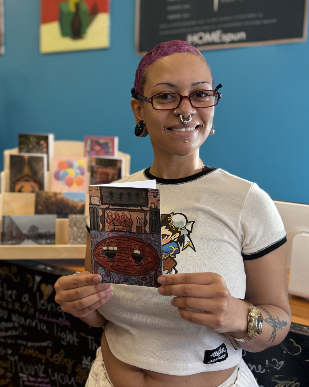 Woman with purple hair holds up a holiday card with a hand-painted image on its face
