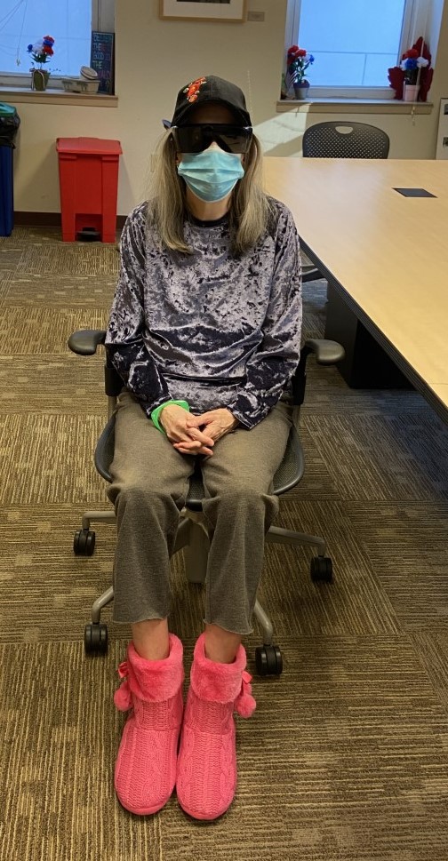 Morgen sitting on a chair at Project HOME's Kate's Place in Center City. She is wearing pink slippers, a blue surgical mask, and dark sunglasses.