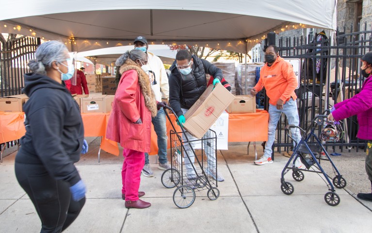 Members of the Project HOME community gather in front of tables full of boxes packed with turkeys and fixings for Thanksgiving. They help their neighbors load up and get their boxes home safely. 
