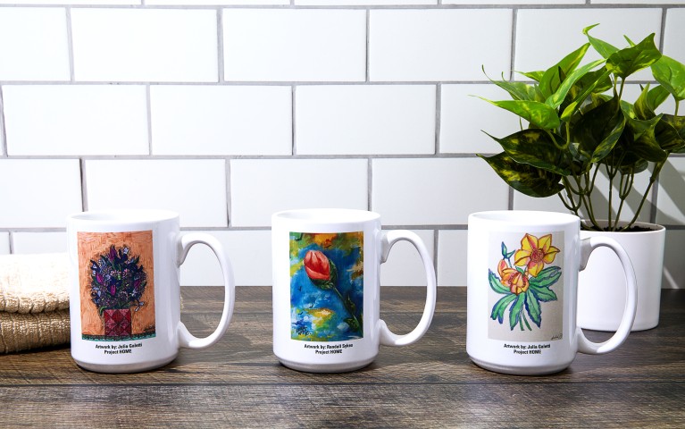 Three mugs with artistic paintings on a kitchen table with a plant behind.