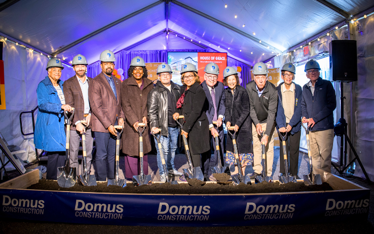 Group of people standing in front of a box of dirt holding shovels and wearing hard hats.