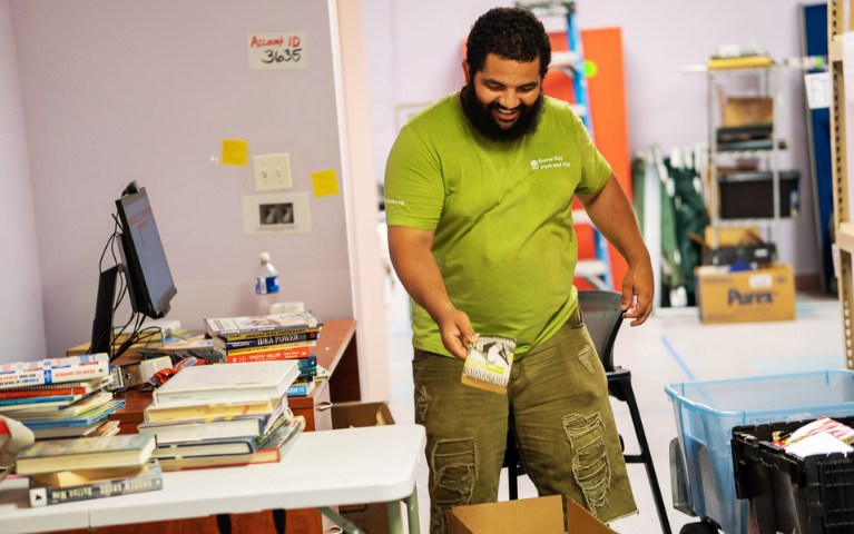 A man in a green shirt and khaki shorts sorts books into boxes.