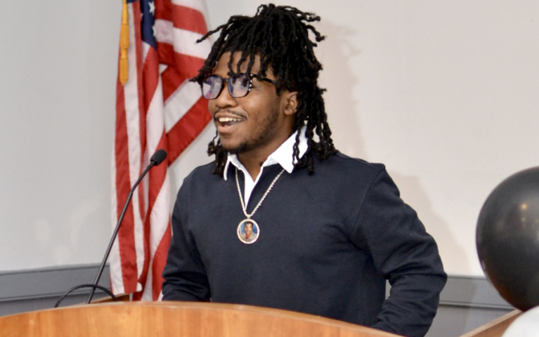 Hamin Muhammad smiles while speaking at a podium in front of an American flag at Project HOME's Honickman Learning Center Comcast Technology Labs
