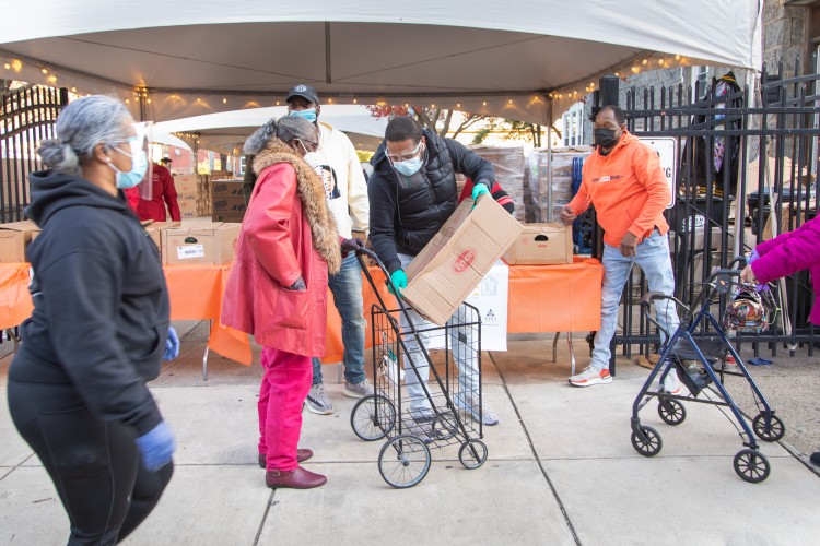 Members of the Project HOME community gather in front of tables full of boxes packed with turkeys and fixings for Thanksgiving. They help their neighbors load up and get their boxes home safely. 