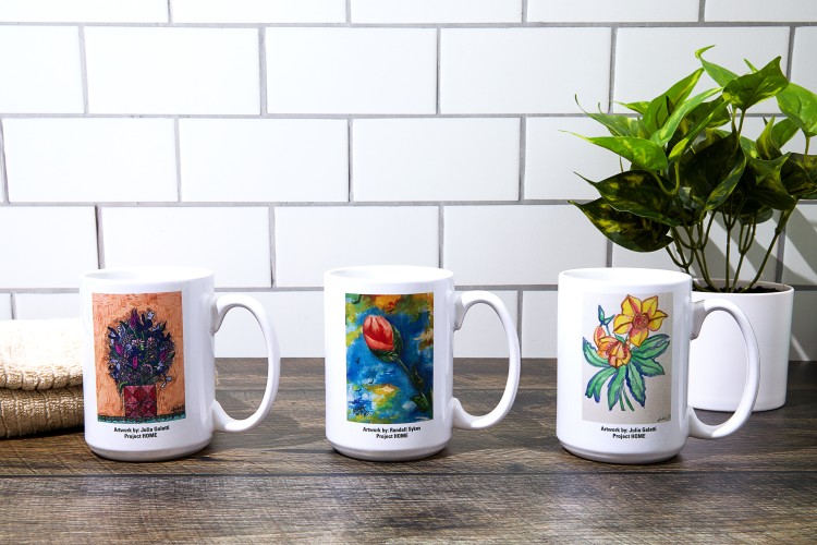 Three mugs with artistic paintings on a kitchen table with a plant behind.