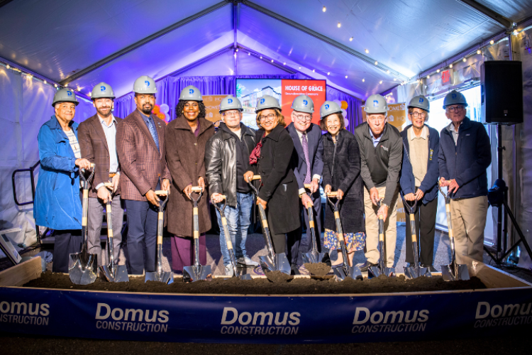 Group of people standing in front of a box of dirt holding shovels and wearing hard hats.
