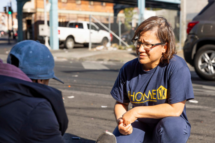 A woman crouching and speaking to another woman sitting on sidewalk