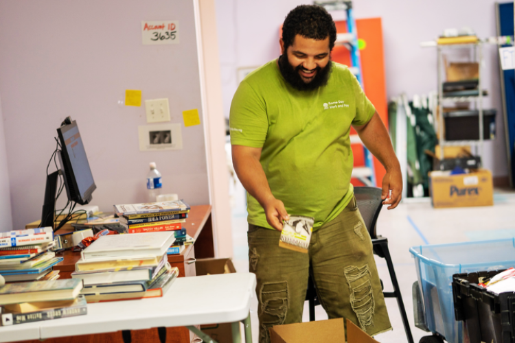 A man in a green shirt and khaki shorts sorts books into boxes.
